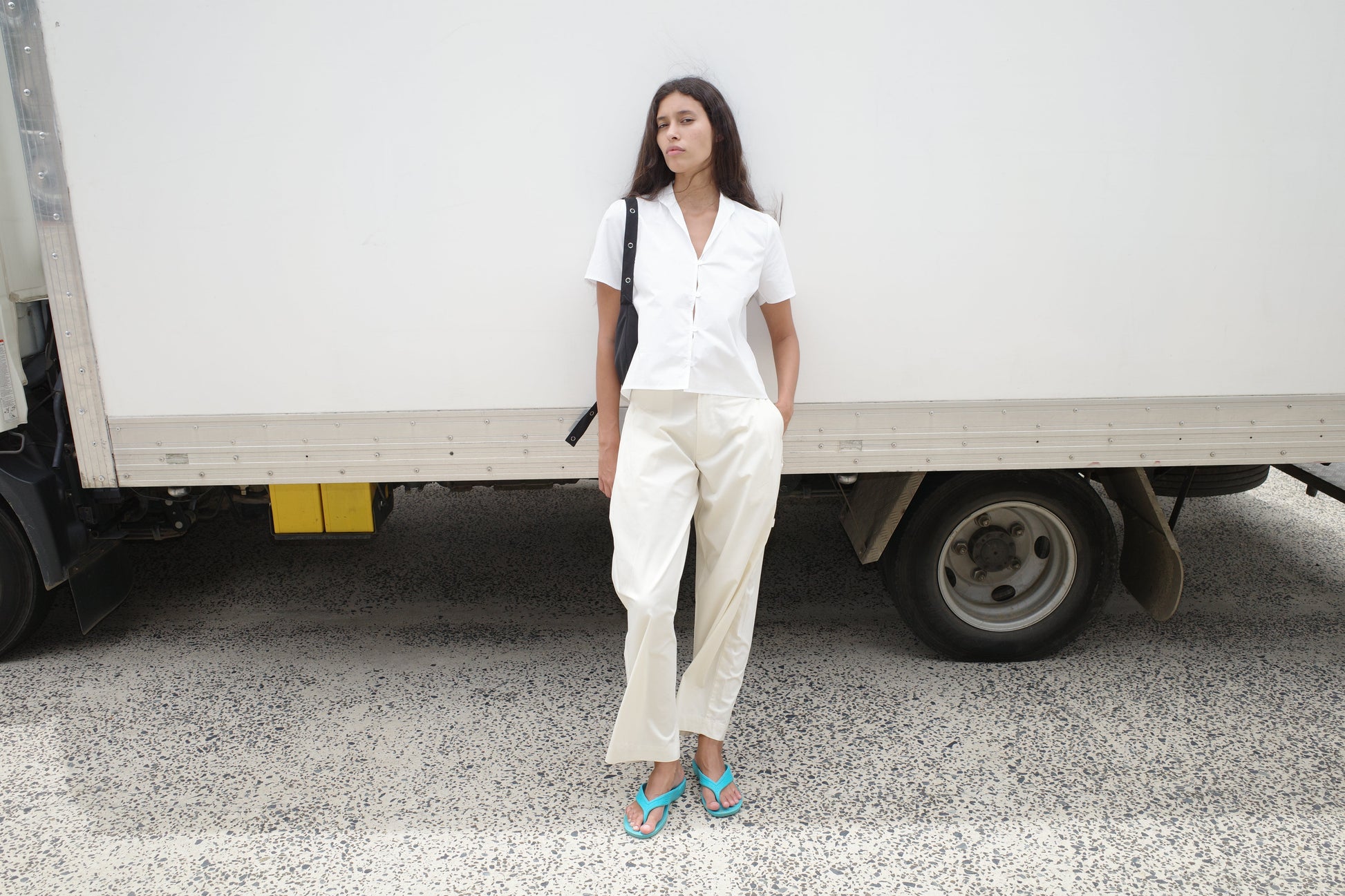 A woman in the Sails Top - White and loose white pants stands by a white truck, one hand in her pocket, looking to the side. She has long dark hair, turquoise flip-flops, and carries a black shoulder bag.
