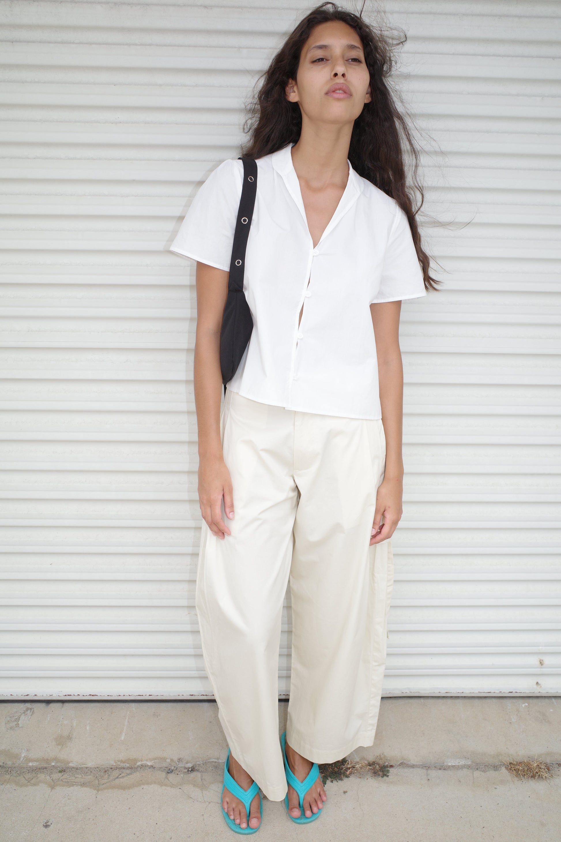 A woman with long dark hair stands before a white corrugated metal door. She wears the Sails Top in White by Deiji Studios, loose cream-colored pants, turquoise sandals, and carries a black shoulder bag.