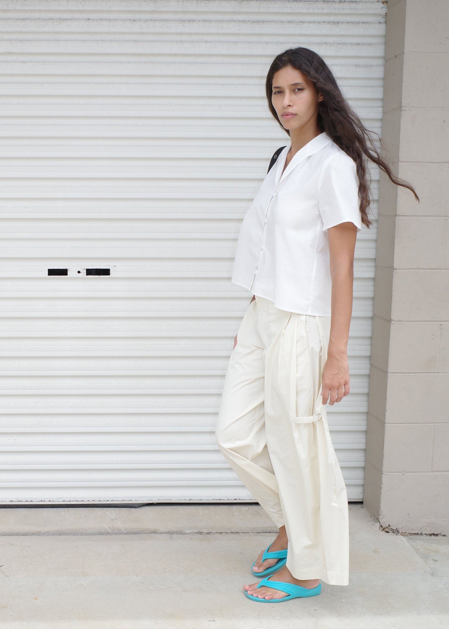 A person with long brown hair wears the Sails Top - White by Deiji Studios, loose cream pants, and teal flip-flops, standing in front of a white metal garage door and looking at the camera.