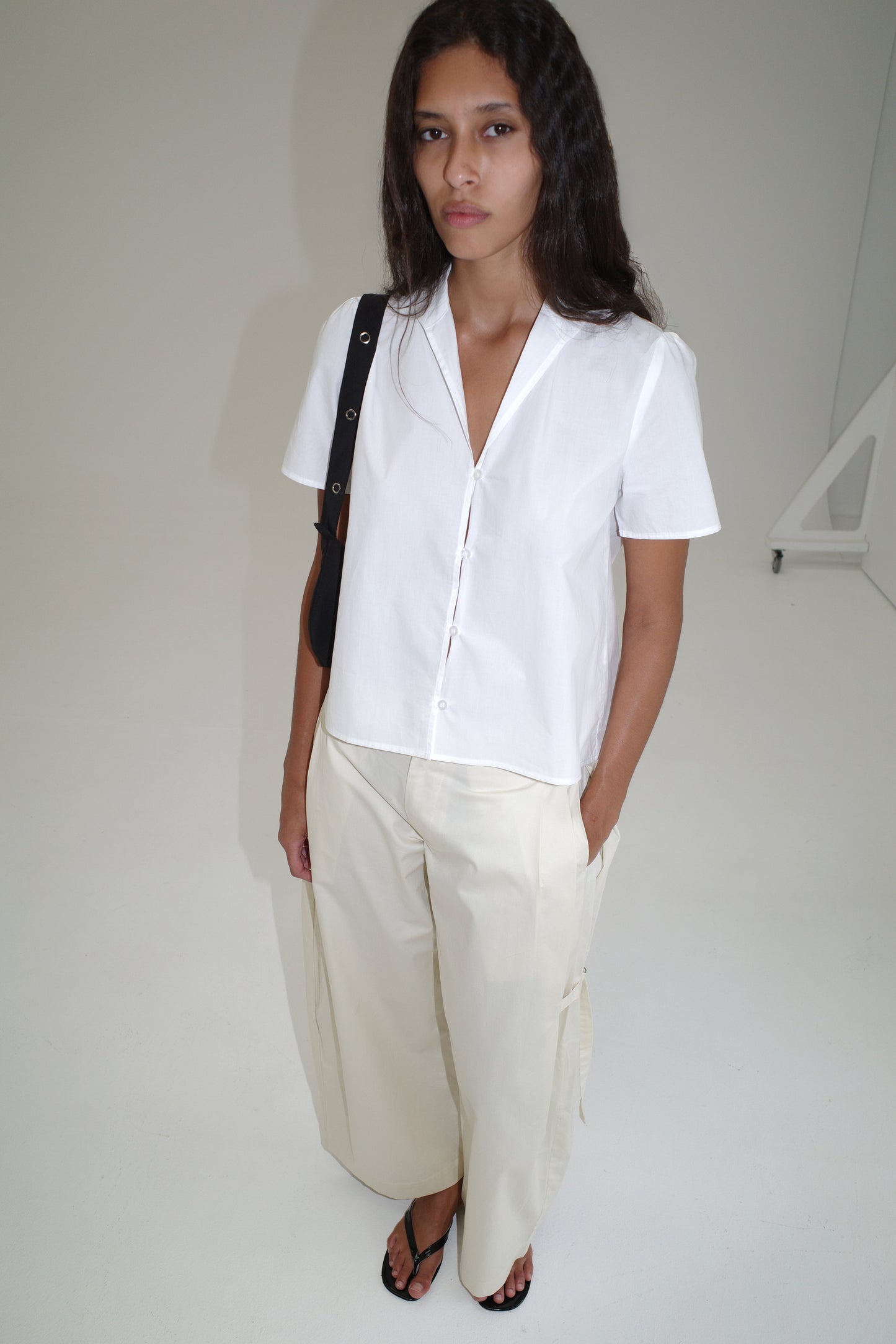 A woman with long dark hair wears the oversized Sails Top - White by Deiji Studios, wide-leg cream pants, black sandals, and a black shoulder bag while standing in a minimalist white room.