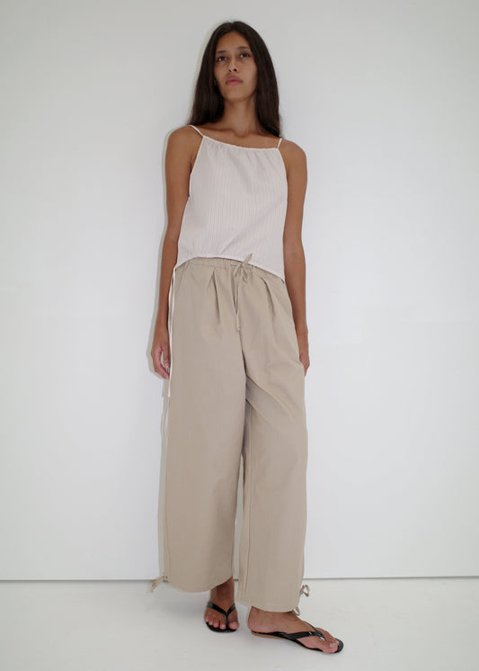 A woman with long, straight hair and a relaxed posture stands against a white background in the Drawcord Wide Leg Pant - Cream by Deiji Studios, paired with a sleeveless top and black flip-flops.
