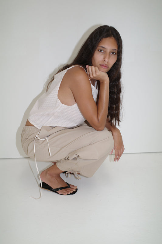 A person with long, wavy hair crouches by a white wall, wearing a Deiji Studios white sleeveless top and the Drawcord Wide Leg Pant in Cream, paired with black sandals, resting their chin on one hand and looking at the camera.