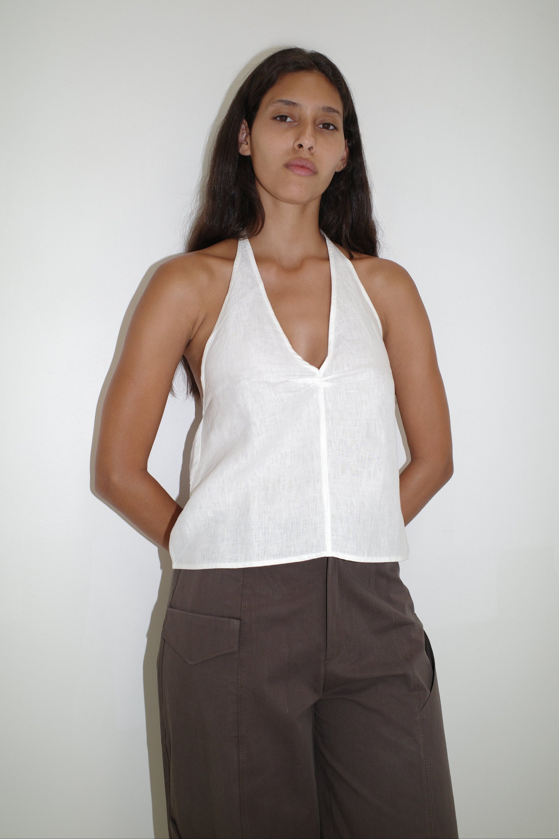 A woman with long dark hair poses confidently against a white background, wearing the sleeveless white Linen Halter Top and high-waisted brown pants, her arms relaxed at her sides.