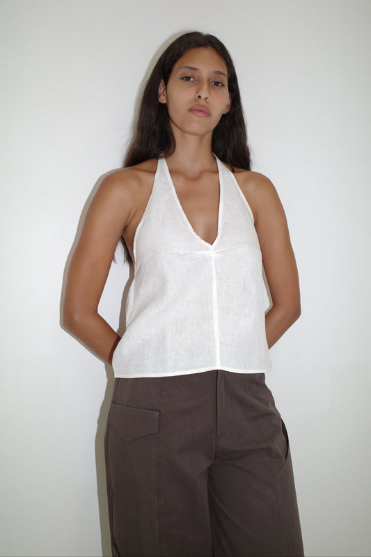 A woman with long dark hair poses confidently against a white background, wearing the sleeveless white Linen Halter Top and high-waisted brown pants, her arms relaxed at her sides.