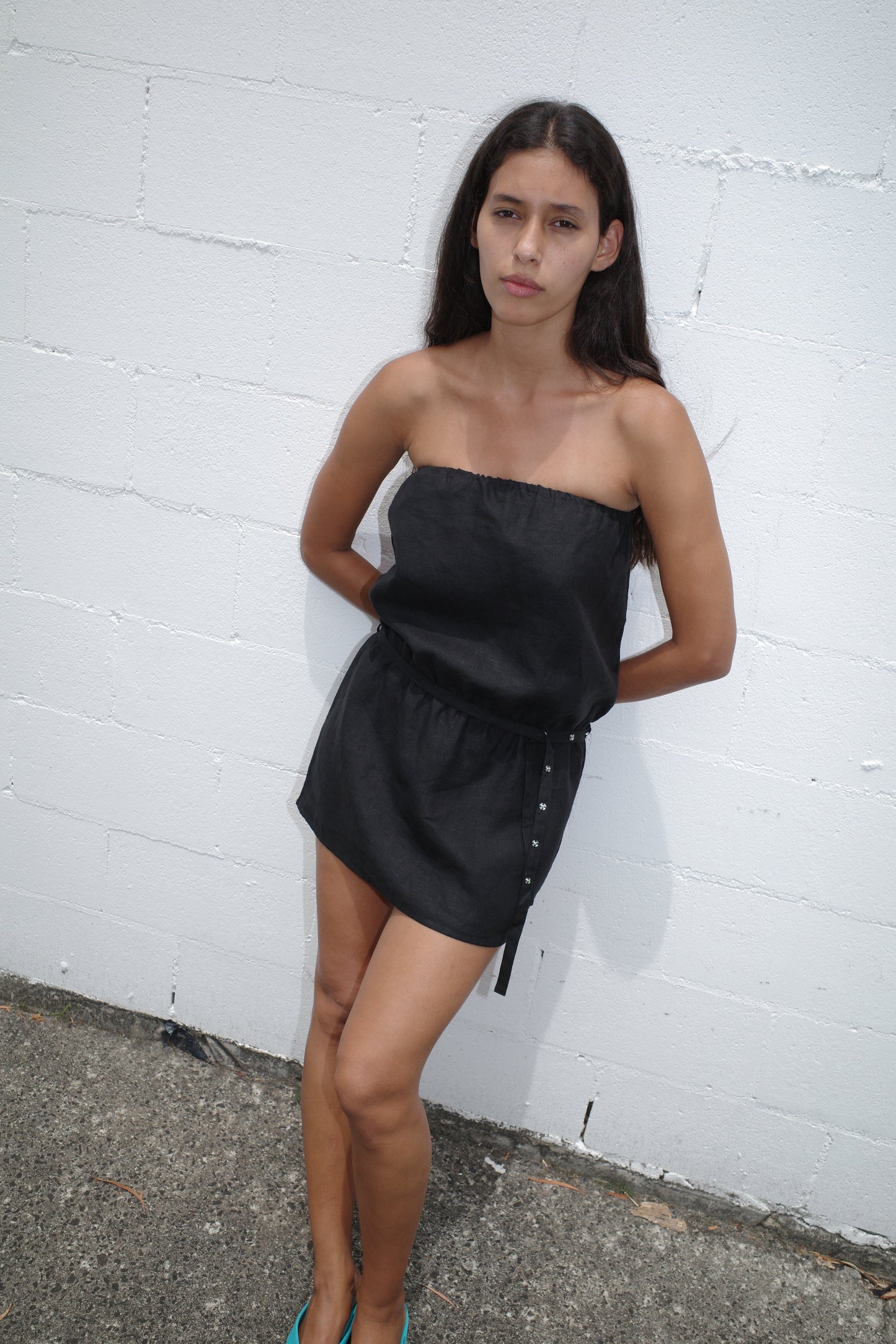A woman with long dark hair stands before a white brick wall in a Strapless Linen Dress - Black, her hands behind her back and a serious expression on her face.
