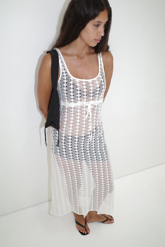 A woman with long brown hair stands against a white background, wearing the Thread Dress - Off White over a black bikini, paired with black sandals and a black shoulder bag.
