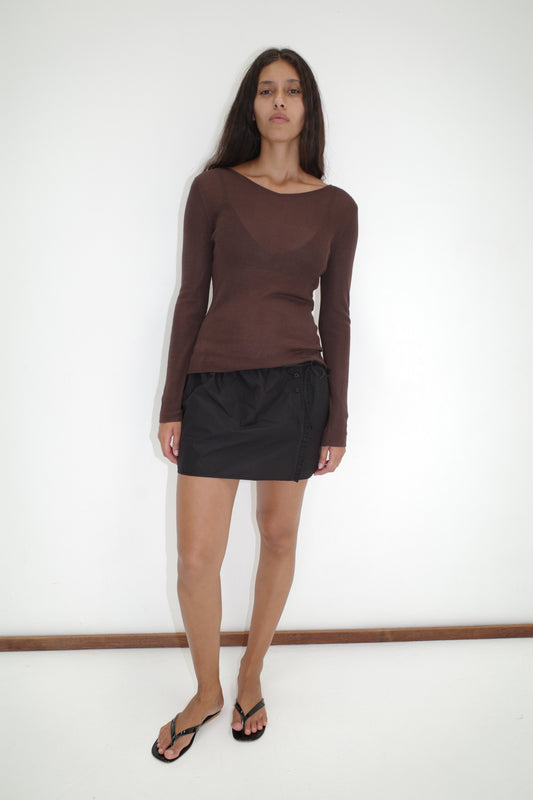 A woman with long dark hair stands against a white wall, wearing the Knitted Top - Dark Brown from Deiji Studios, a black mini skirt, and black flip-flops. She gazes at the camera with a neutral expression.