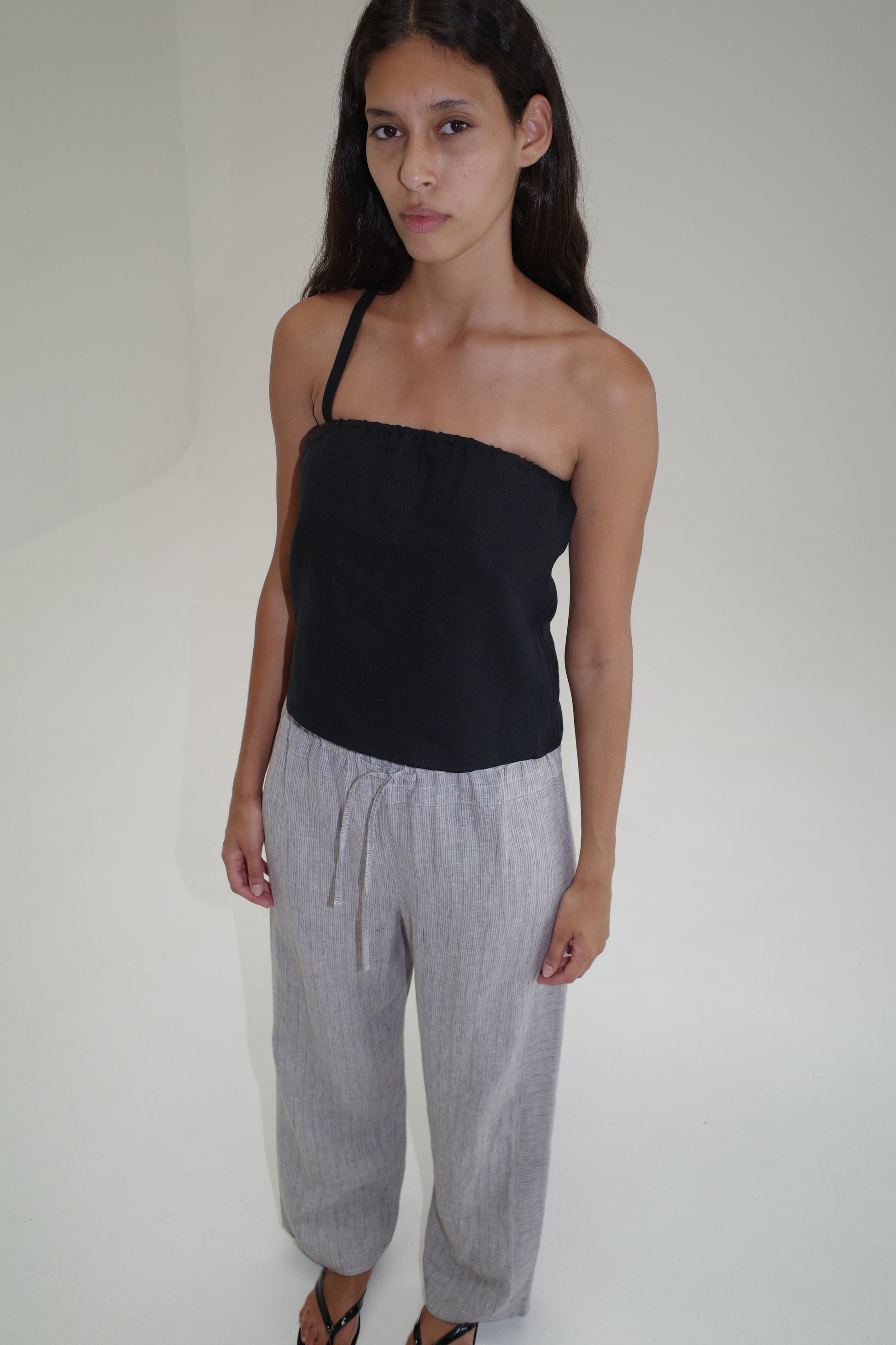 A woman with long dark hair stands in a minimalist, light-colored room, wearing a black strapless top and the Double Waist Linen Pant in Haze Blue, crafted from organic French linen. She gazes slightly away from the camera with a neutral expression.