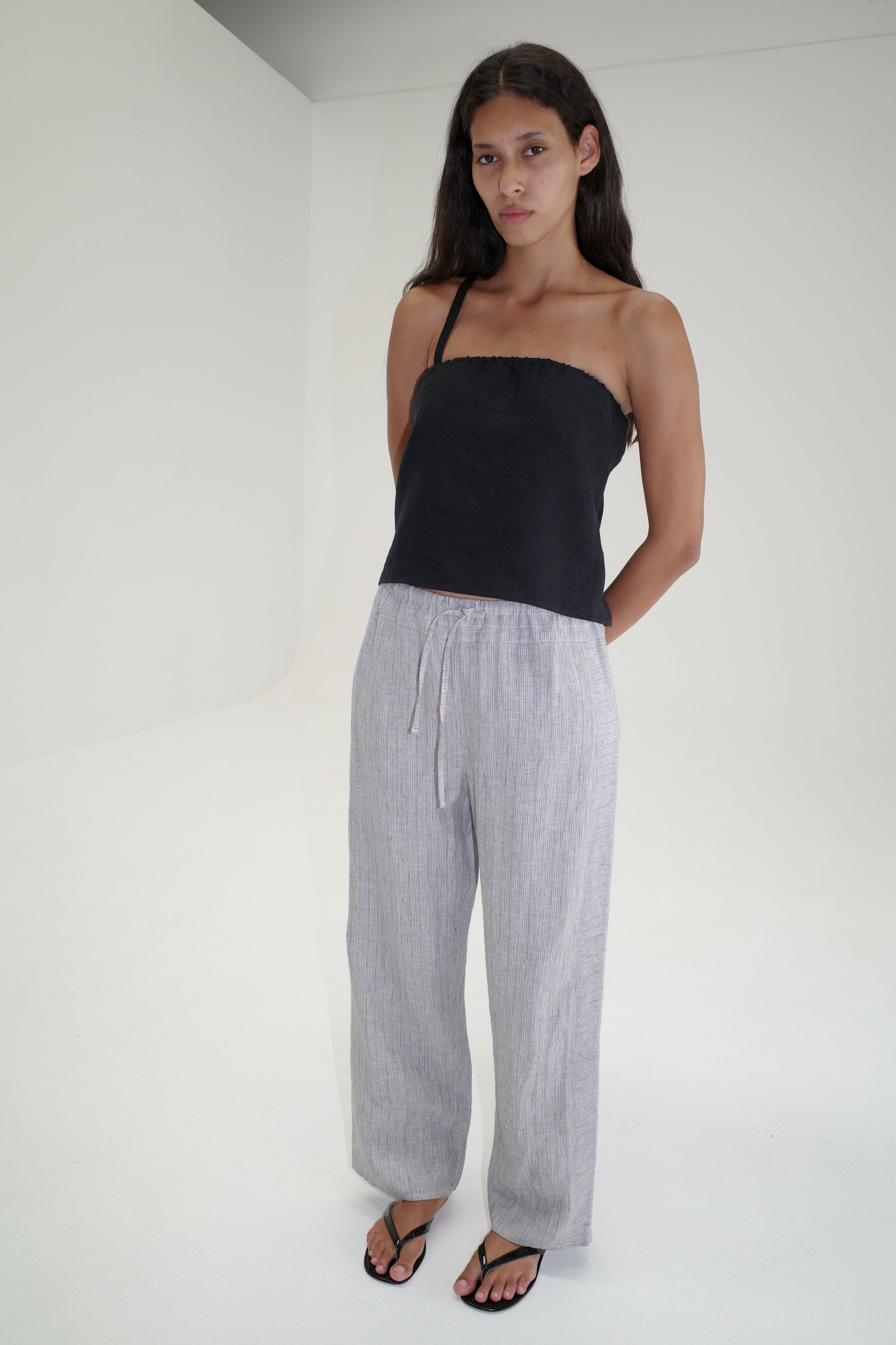 A person with long brown hair stands in a minimal white room, wearing a black strapless top, Double Waist Linen Pant - Haze Blue, and black sandals. Their arms are relaxed behind their back.
