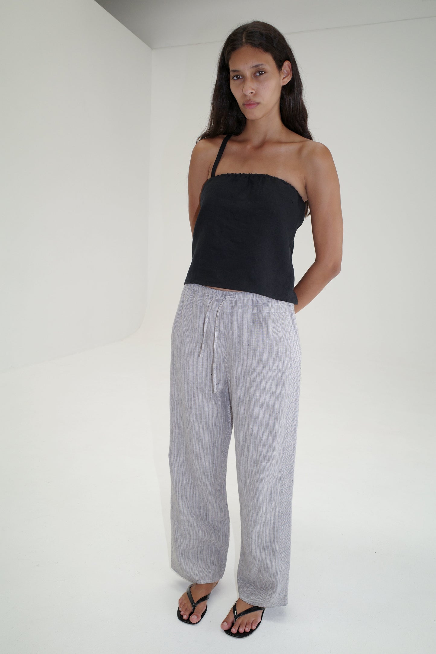 A woman with long dark hair stands in a minimal white room, wearing a black strapless top, Deiji Studios Double Waist Linen Pant in Haze Blue organic French linen, and black sandals. Her hands are behind her back and her expression is neutral.