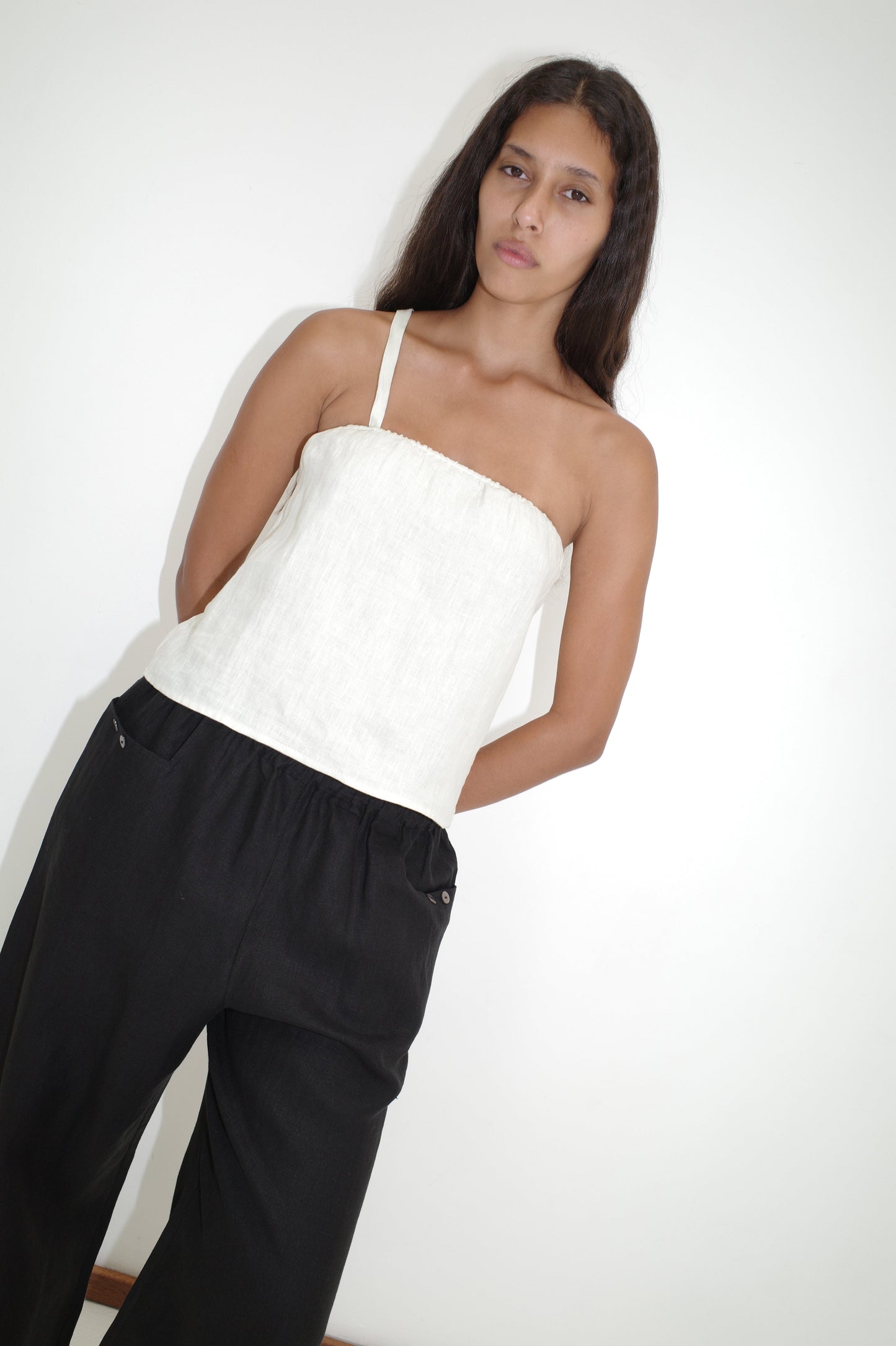 A woman with long dark hair stands against a white wall, wearing the Button Strap Top - White by Deiji Studios and loose black pants. She gazes calmly at the camera with her arms behind her back.