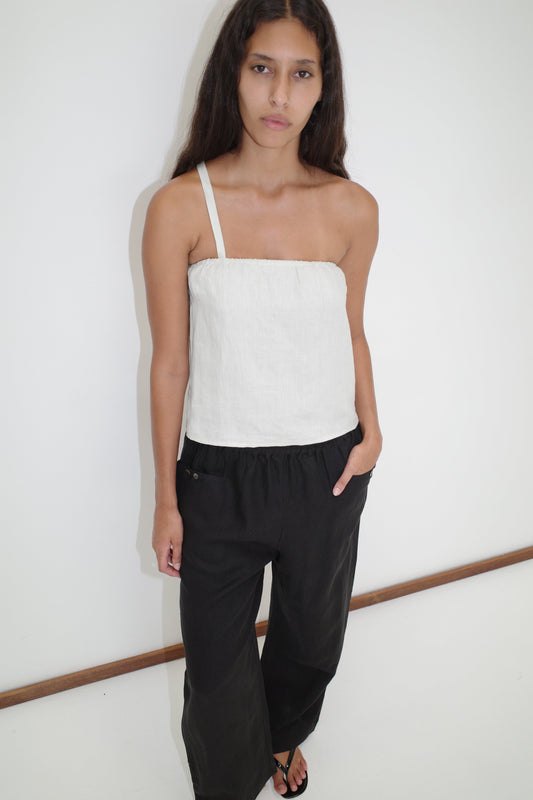 A woman with long dark hair wears the Button Strap Top - White by Deiji Studios in French linen and loose black pants, standing against a plain white wall with one hand in her pocket.