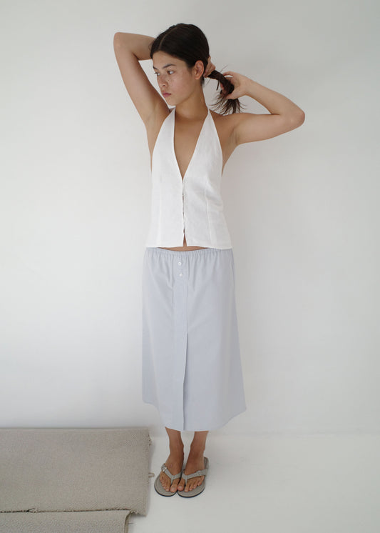 A person with dark hair stands against a white wall, adjusting their hair while wearing a sleeveless white halter top, flip-flops, and the Overlap Skirt in light gray that falls to the knee.