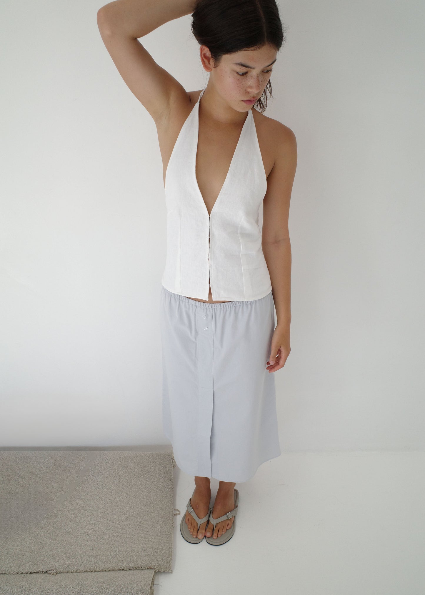 A woman barefoot on a white floor stands against a plain white background, wearing a sleeveless white halter top and the Overlap Skirt - Blue, looking down with one hand raised to her head.