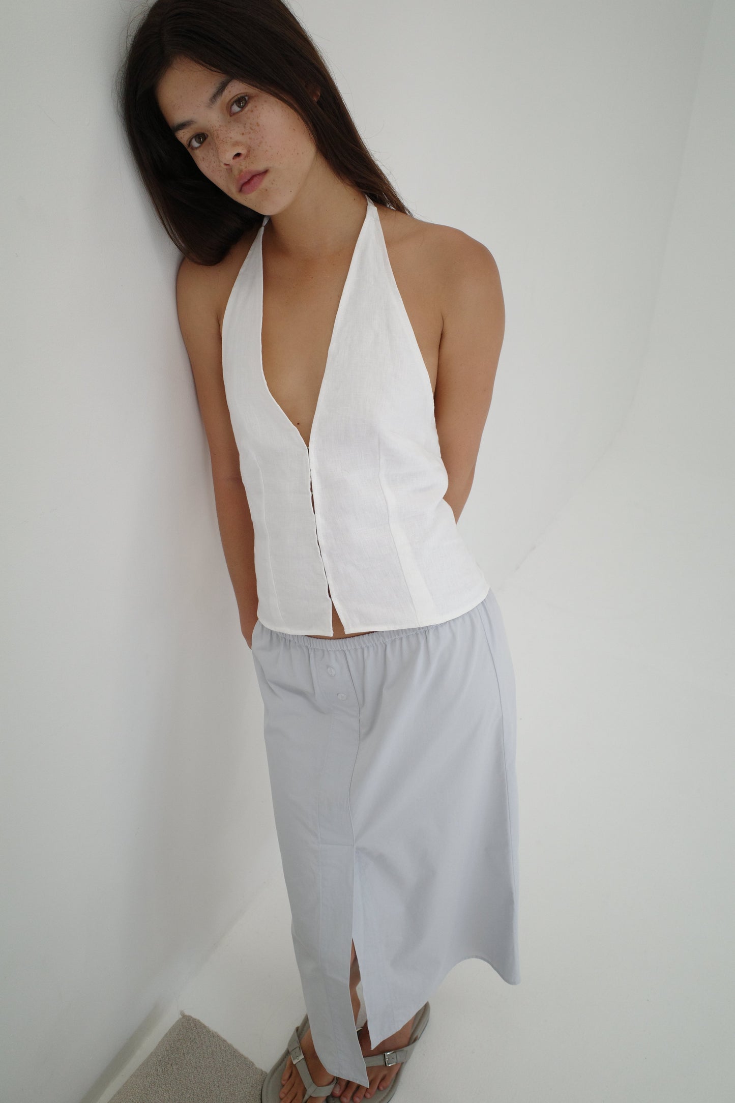 A woman with long dark hair stands against a white wall, wearing a sleeveless white halter top and the Overlap Skirt - Blue by Deiji Studios. The mid-length skirt features a front slit. She looks up slightly, hands behind her back, and wears sandals.