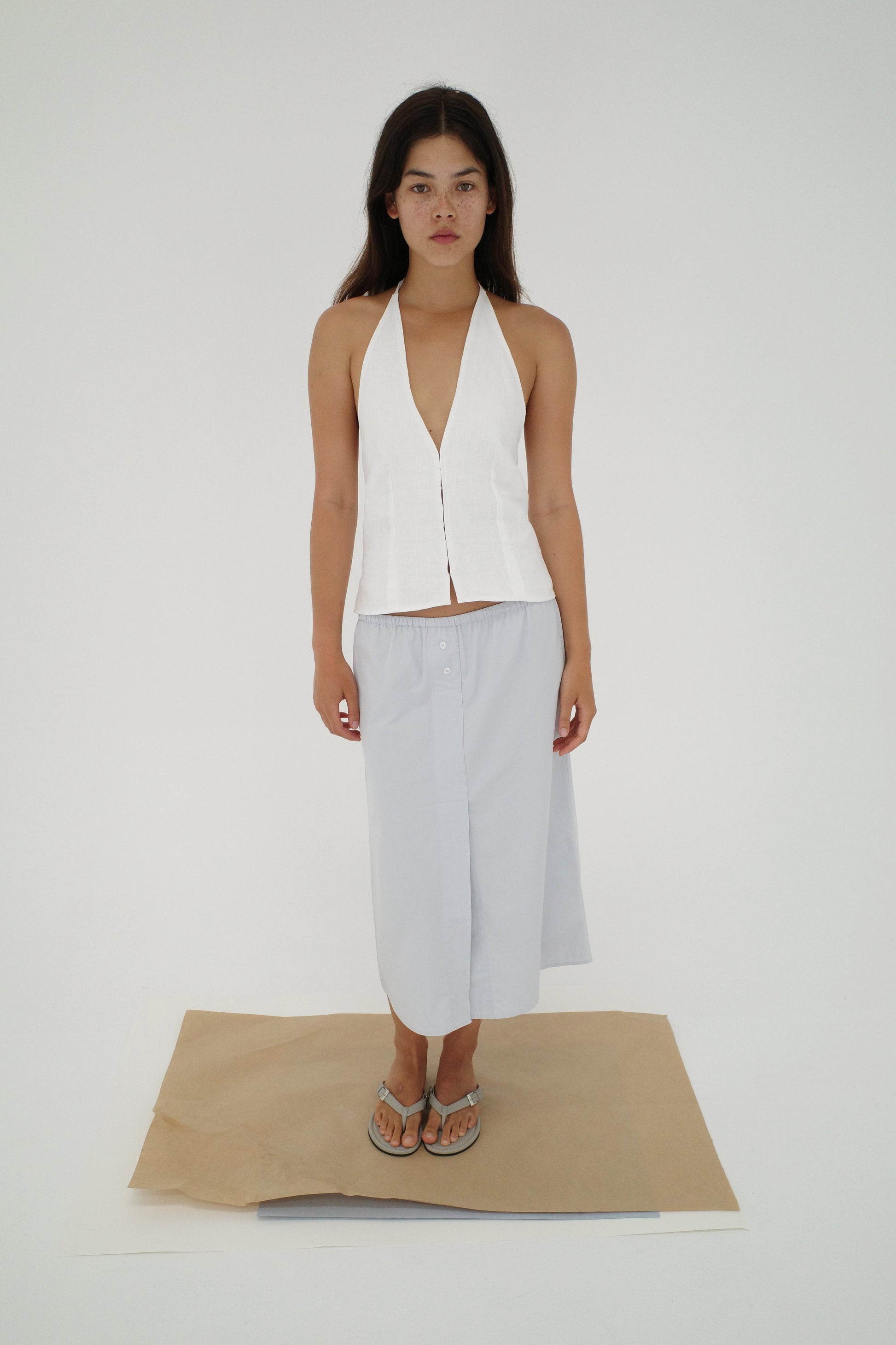 A woman stands on brown and white paper in a white room, wearing a sleeveless white halter top, the Overlap Skirt - Blue by Deiji Studios, and flip-flops. She faces forward with a neutral expression.