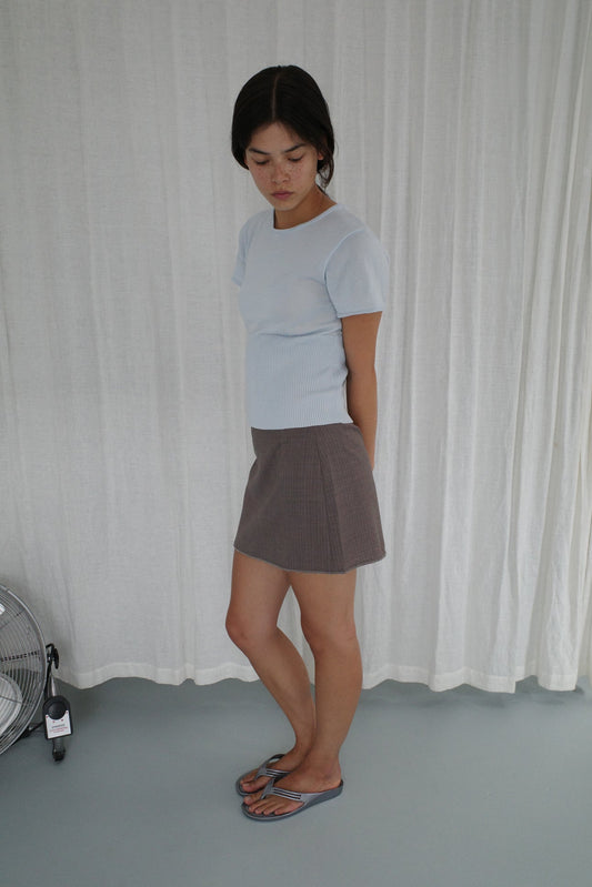 A woman stands indoors against white curtains, looking down with her hands behind her back. She wears the Trim Knit Tee - Ice Blue from Deiji Studios, a gray mini skirt, and gray sandals. A small fan sits on the floor nearby.