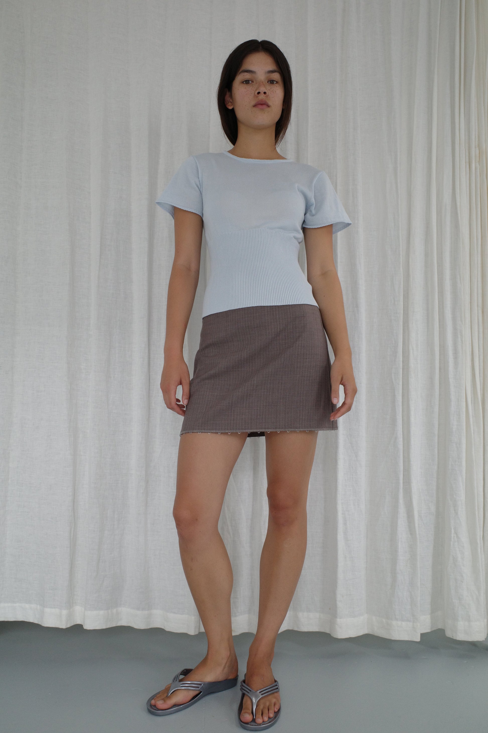 A woman stands before white curtains, confidently facing the camera in Deiji Studios’ Trim Knit Tee - Ice Blue, paired with a brown mini skirt and blue sandals.