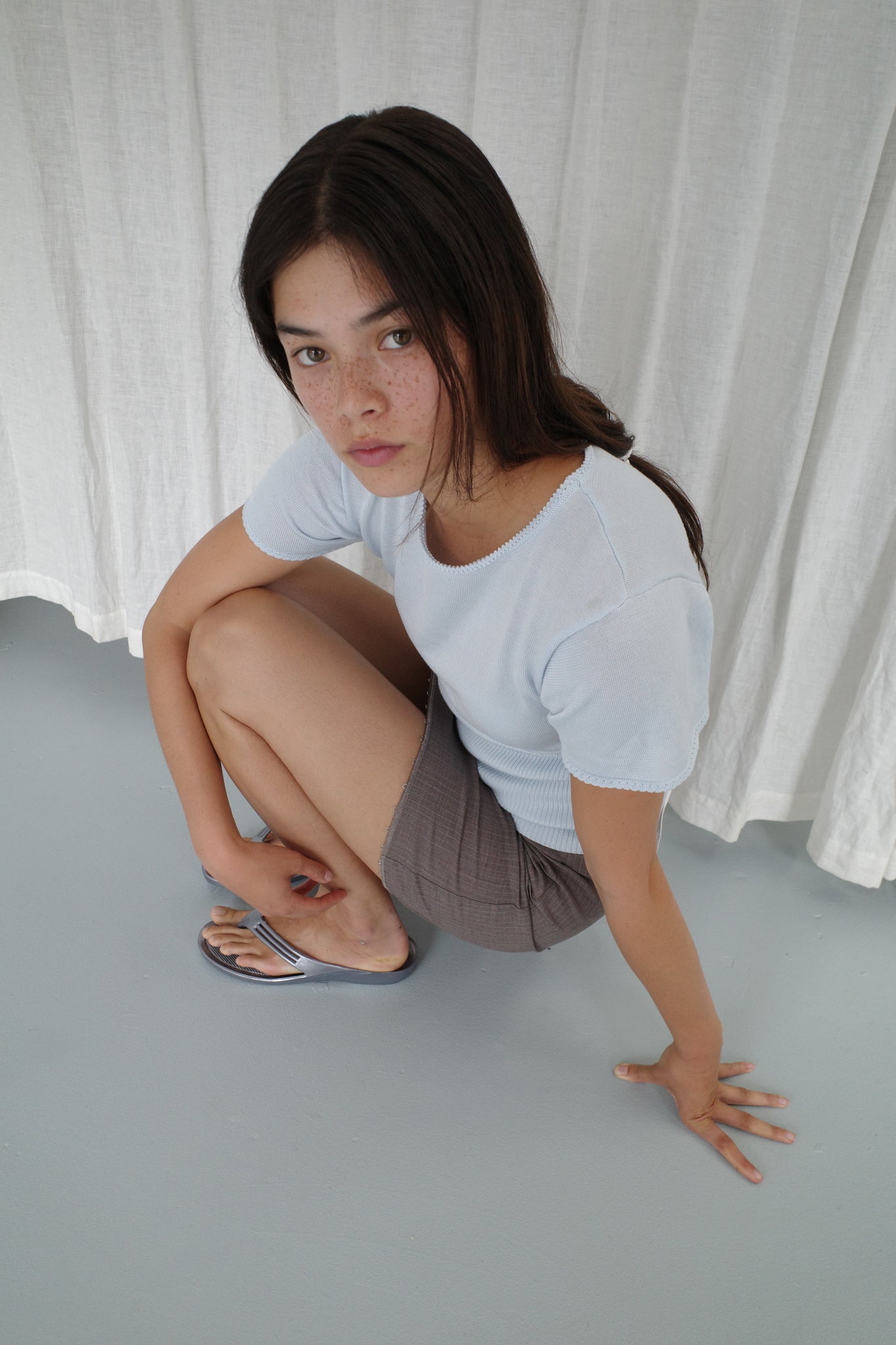 A young woman with long dark hair and freckles crouches on a light gray floor, wearing Deiji Studios’ Trim Knit Tee in Ice Blue, brown shorts, and silver sandals. She looks at the camera with a white curtain behind her.