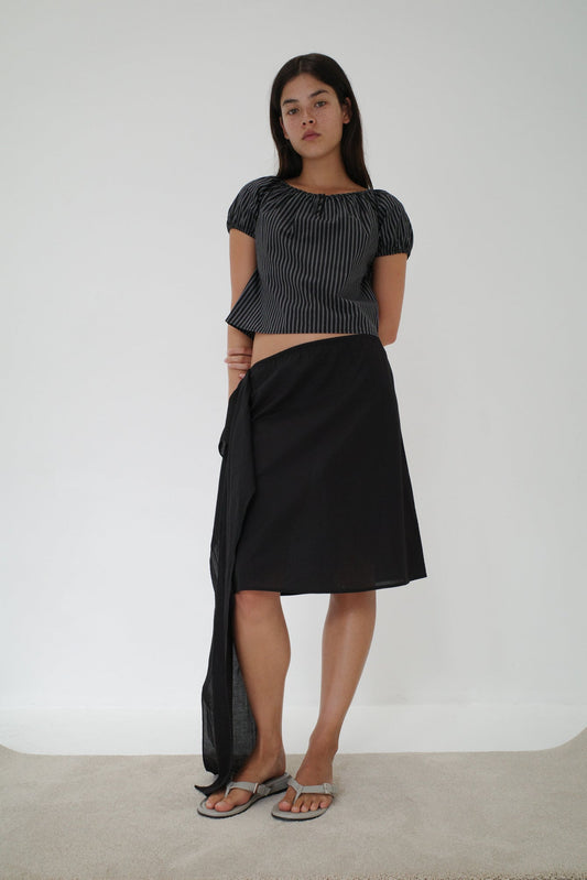 A woman with long dark hair and a neutral expression stands indoors against a plain white wall, wearing a black-and-grey striped top, the Scarf Midi - Black skirt from Deiji Studios with an elasticated waistband, and sandals.