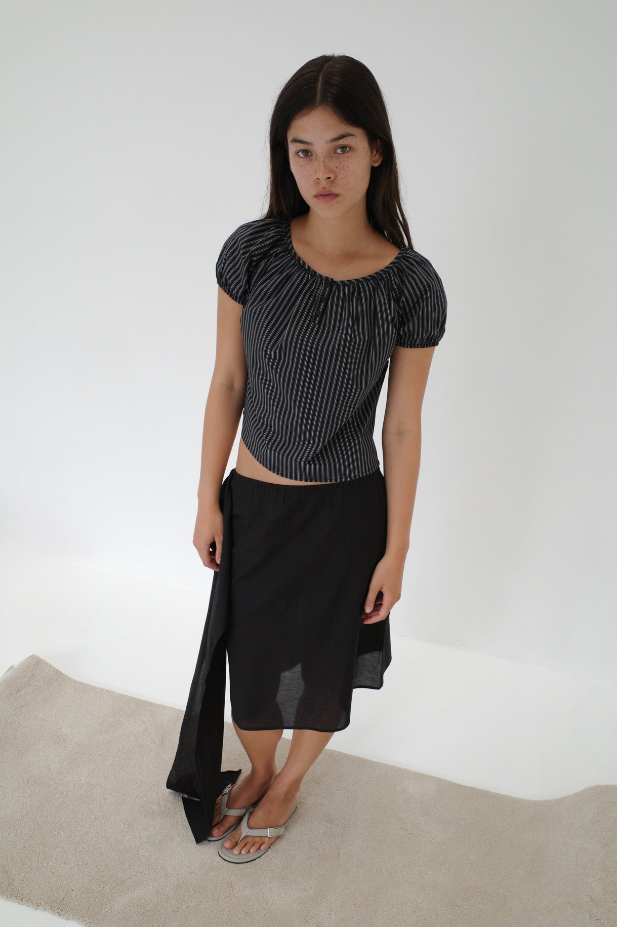 A young woman with long brown hair stands on a light rug against a white background, wearing the Scarf Midi - Black skirt with an elasticated waistband, a striped top, and flip-flops, looking directly at the camera.