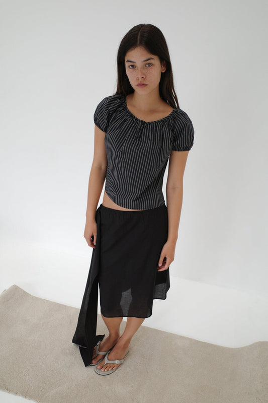 A young woman with long brown hair stands on a light rug against a white background, wearing the Scarf Midi - Black skirt with an elasticated waistband, a striped top, and flip-flops, looking directly at the camera.