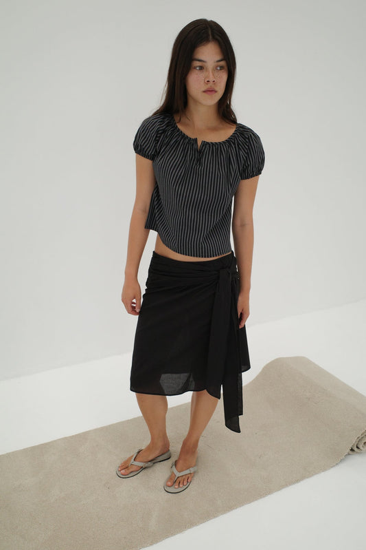 A woman with long dark hair stands on a light rug in a minimal studio, wearing the Scarf Midi - Black skirt from Deiji Studios with an elasticated waistband, a striped short-sleeve top, and flip-flops. She looks slightly to the side.