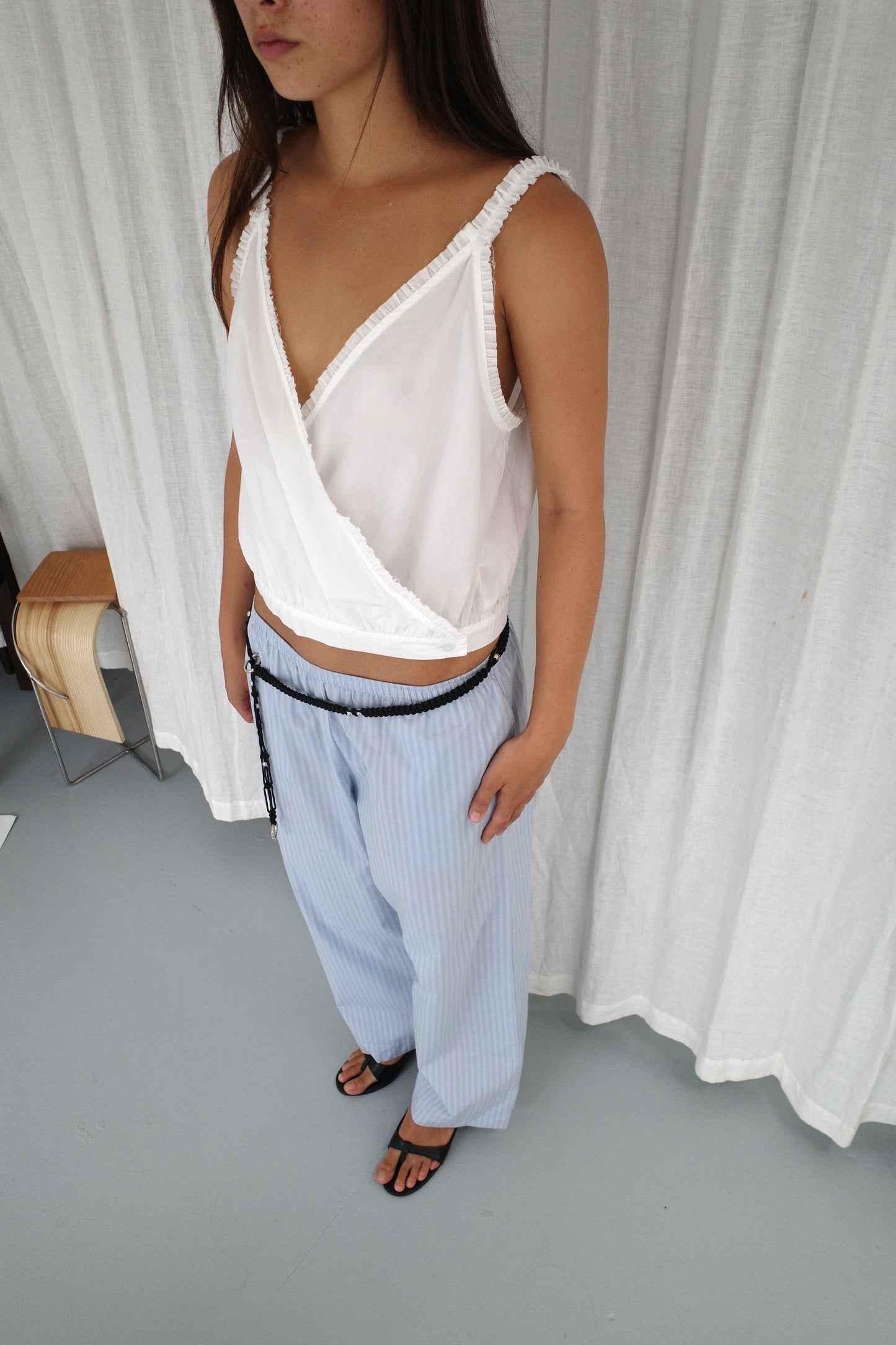 A woman stands indoors wearing the Overlie Top in white, a sleeveless wrap style with a crossover front, paired with loose light blue pinstriped pants and a black belt. She has long brown hair, black sandals, and white curtains in the background.