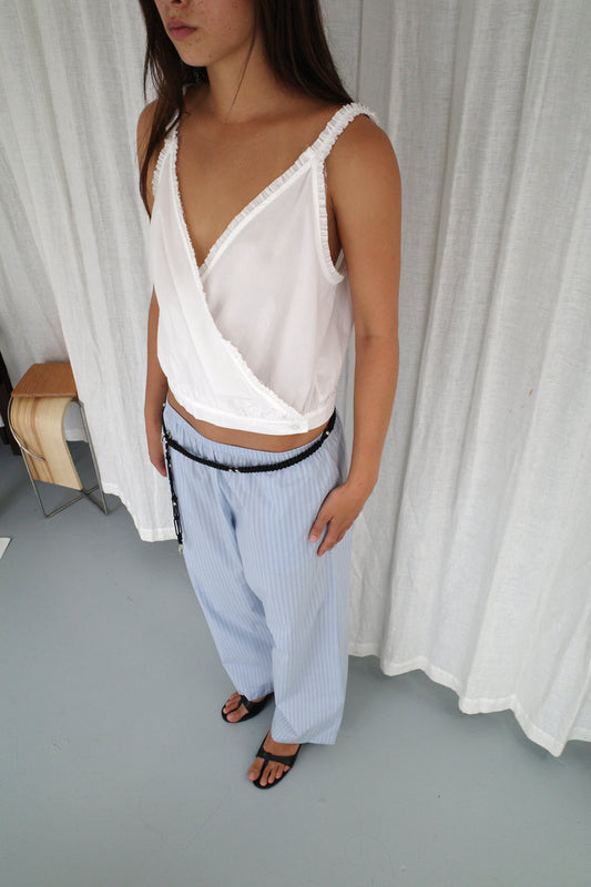 A woman stands indoors wearing the Overlie Top in white, a sleeveless wrap style with a crossover front, paired with loose light blue pinstriped pants and a black belt. She has long brown hair, black sandals, and white curtains in the background.