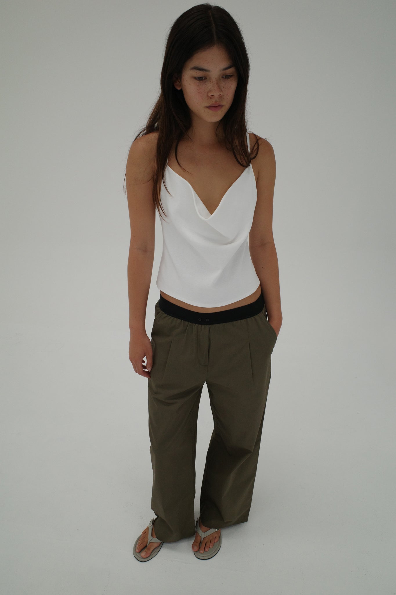 A woman with long brown hair stands against a white background, wearing a white draped tank top, Deiji Studios Contrast Waist Pant - Olive, and flip-flops. She gazes slightly downward with a neutral expression.