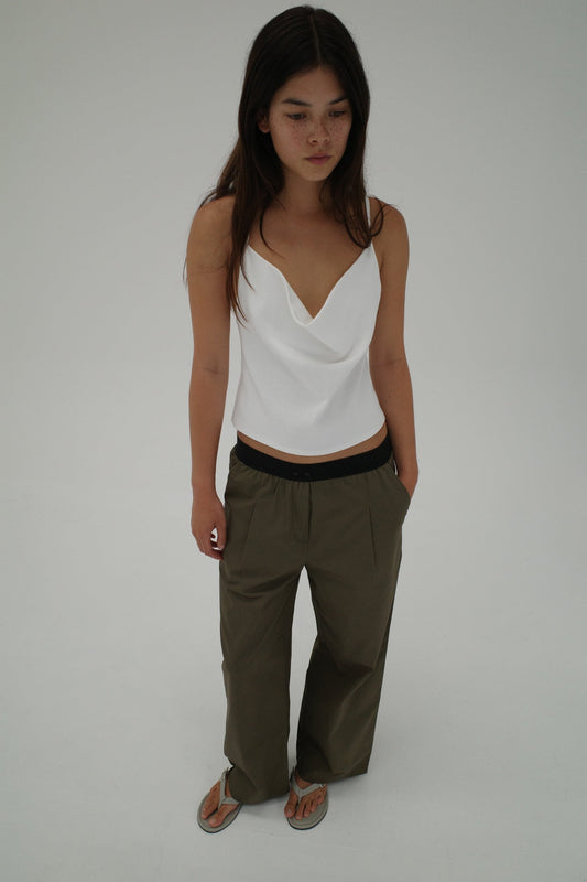 A woman with long brown hair stands against a white background, wearing a white draped tank top, Deiji Studios Contrast Waist Pant - Olive, and flip-flops. She gazes slightly downward with a neutral expression.