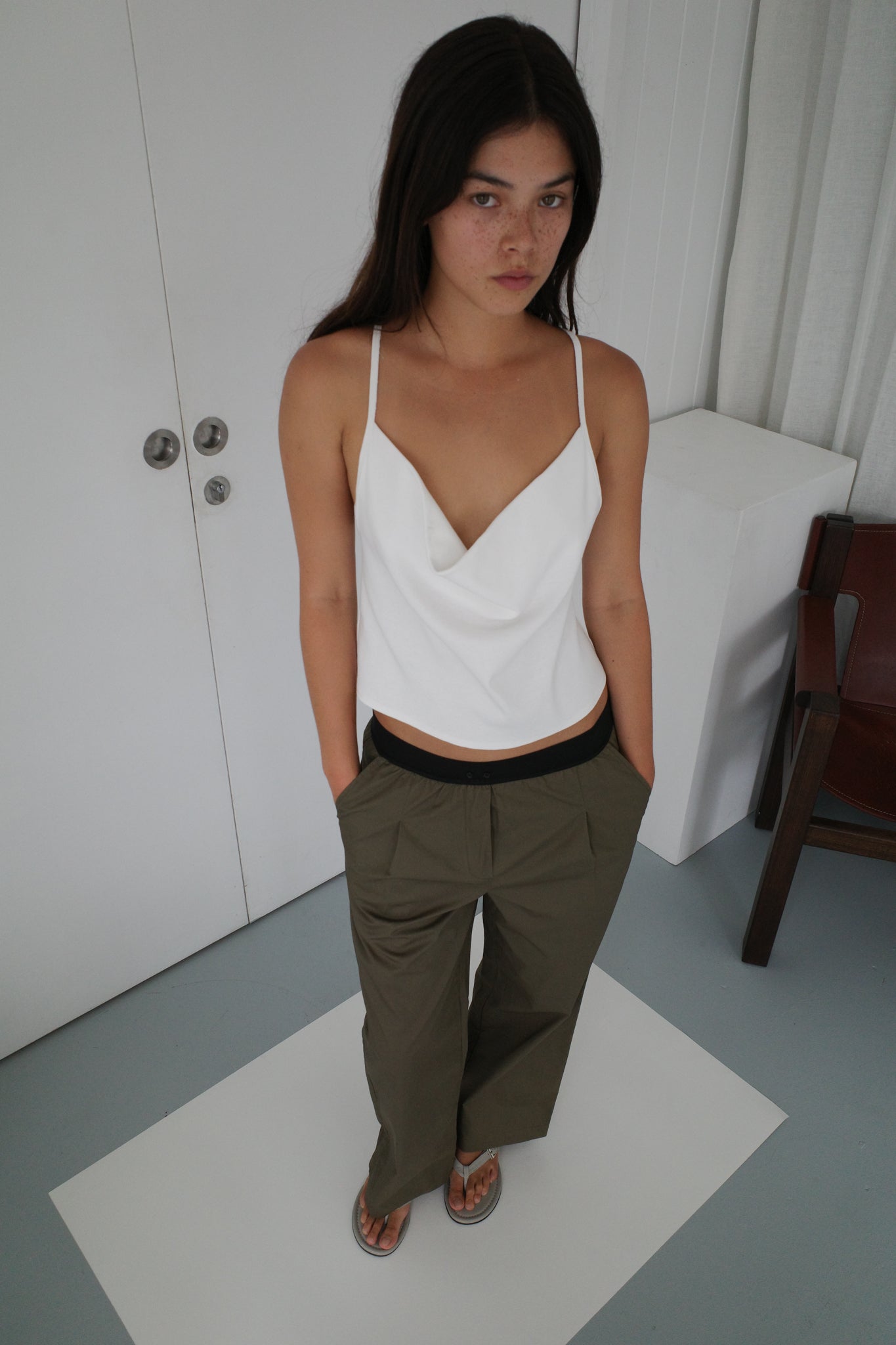 A woman with long dark hair stands indoors, wearing a white sleeveless draped top, Deiji Studios Contrast Waist Pant - Olive in a relaxed straight leg fit, and sandals. Her hands are in her pockets as she looks up at the camera with a neutral expression.