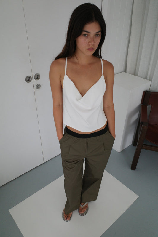 A woman with long dark hair stands indoors, wearing a white sleeveless draped top, Deiji Studios Contrast Waist Pant - Olive in a relaxed straight leg fit, and sandals. Her hands are in her pockets as she looks up at the camera with a neutral expression.