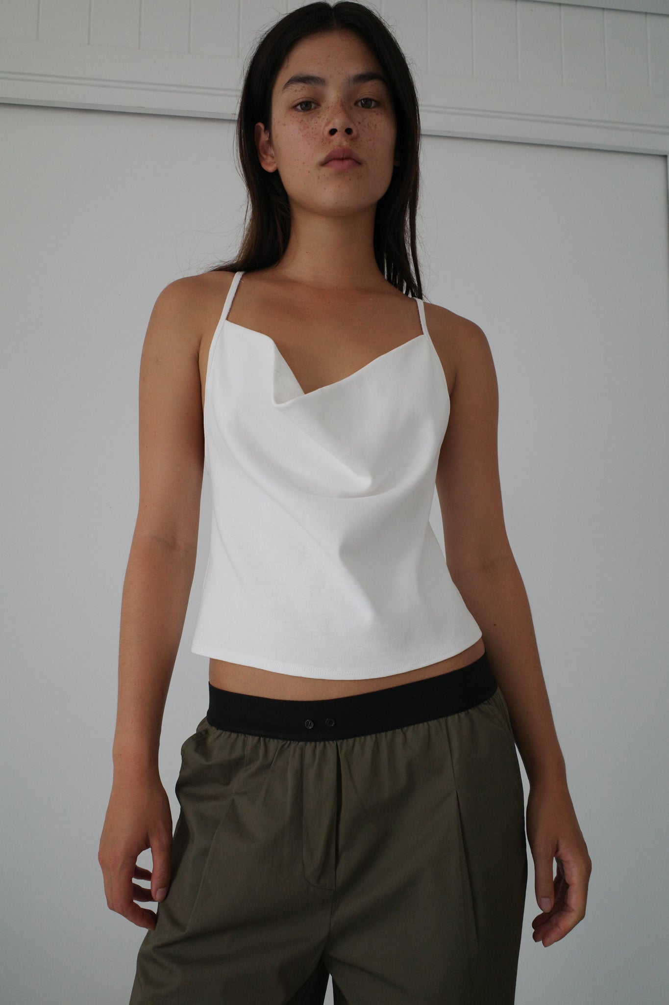A woman with long dark hair stands indoors against a white wall, wearing a white draped camisole and the Olive Contrast Waist Pant from Deiji Studios, looking confidently at the camera.