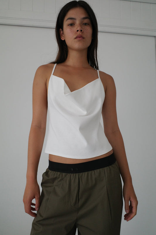 A woman with long dark hair stands indoors against a white wall, wearing a white draped camisole and the Olive Contrast Waist Pant from Deiji Studios, looking confidently at the camera.
