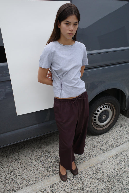 A woman with long brown hair stands in front of a dark van, wearing the Pinch Tee - Grey with a crew neckline, loose dark brown pants, and brown shoes. Her hands are behind her back as she looks at the camera with a neutral expression.
