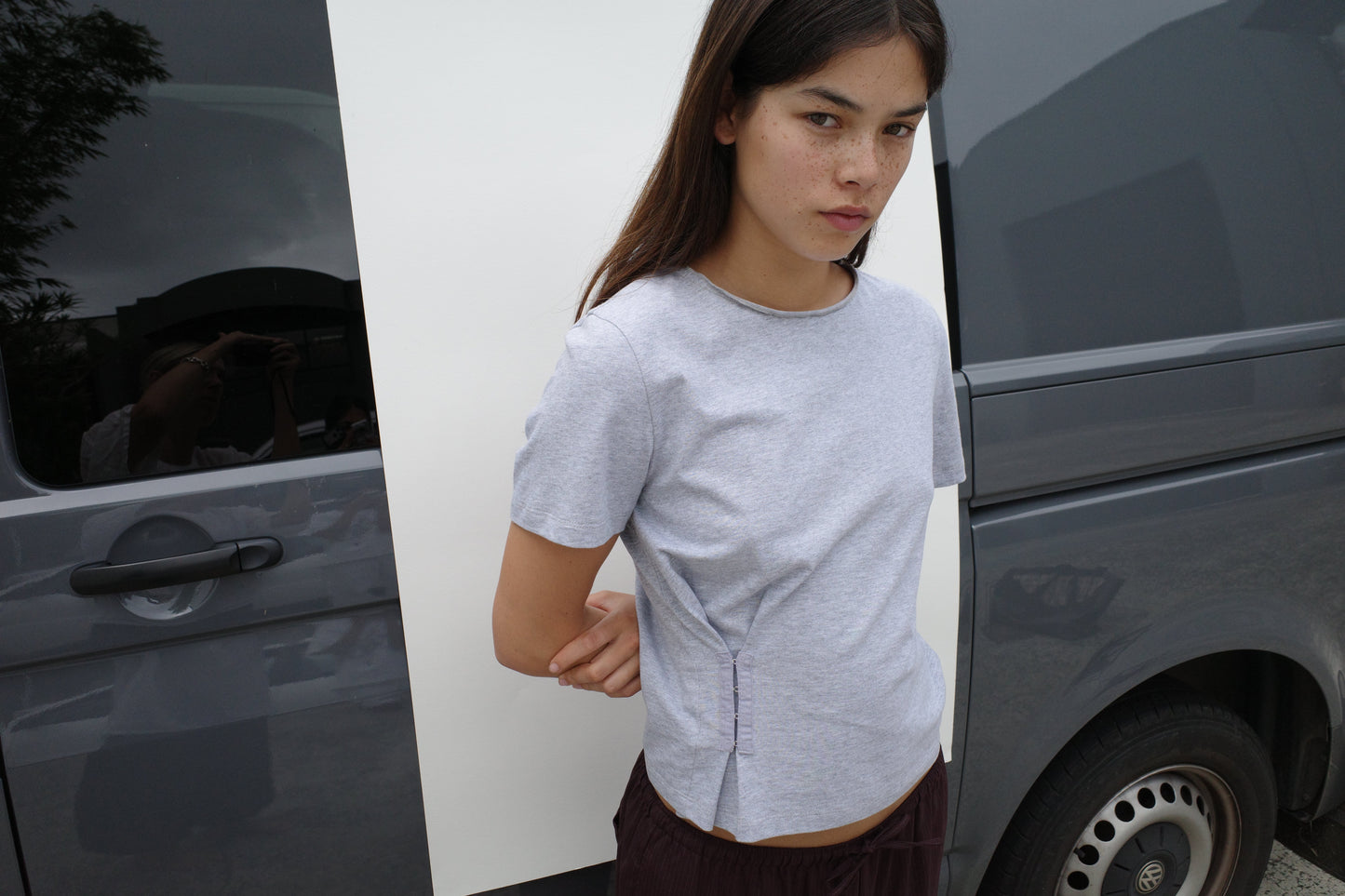 A woman with long dark hair wears the Pinch Tee - Grey and dark pants, standing outdoors in front of a gray van and looking at the camera with a neutral expression.