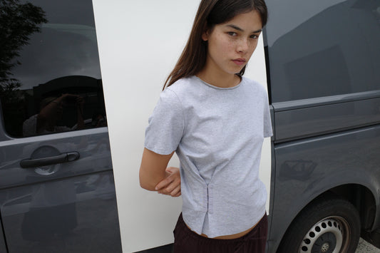 A woman with long brown hair stands in front of a gray van, looking seriously at the camera, wearing the Pinch Tee - Grey with short sleeves and a crew neckline, paired with dark pants. Her arms are crossed behind her back.
