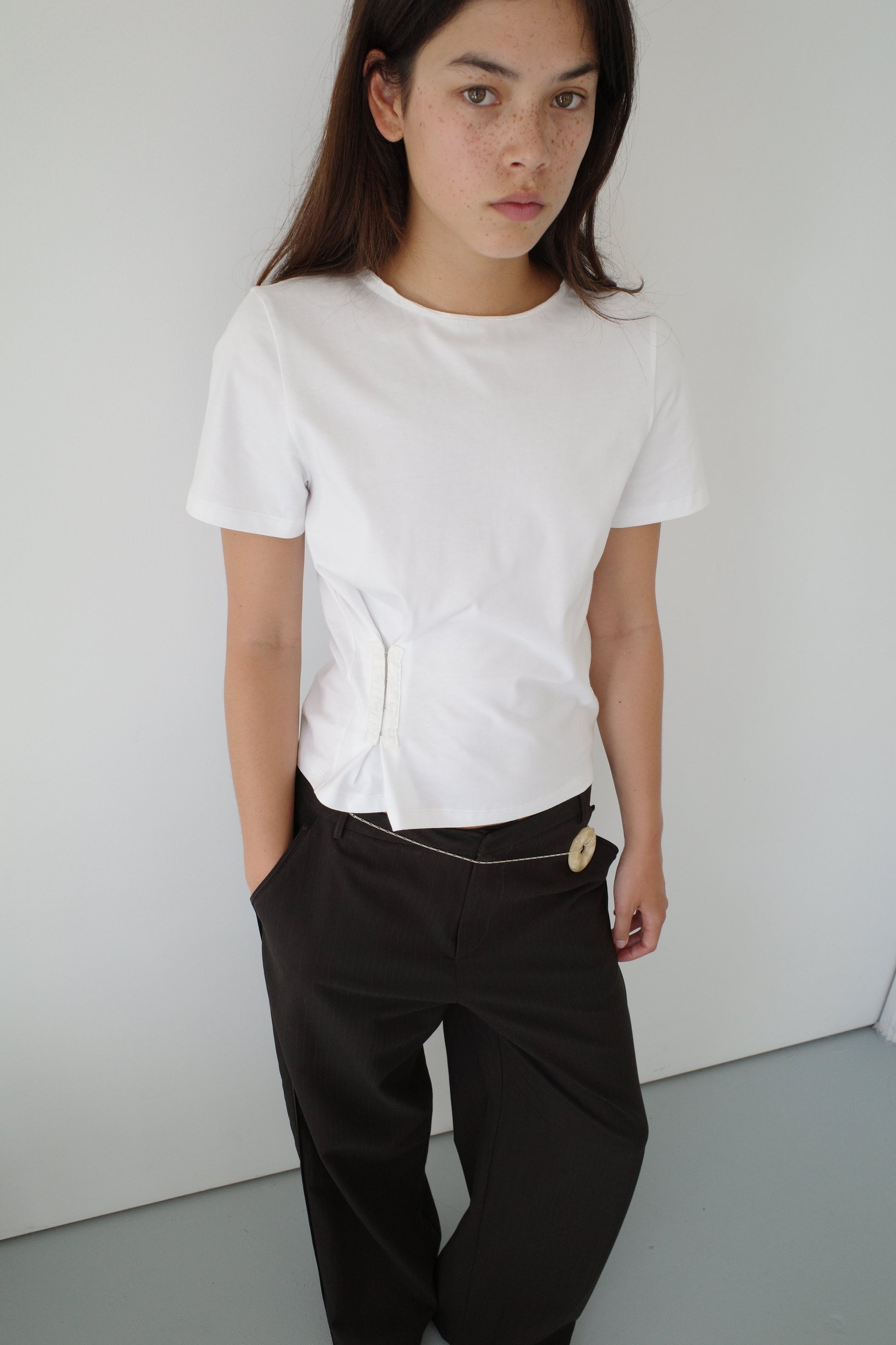 A person with long dark hair wears the Pinch Tee, a white short-sleeve top, paired with loose black pants. They stand against a plain white wall, looking at the camera neutrally with one hand in their pocket.