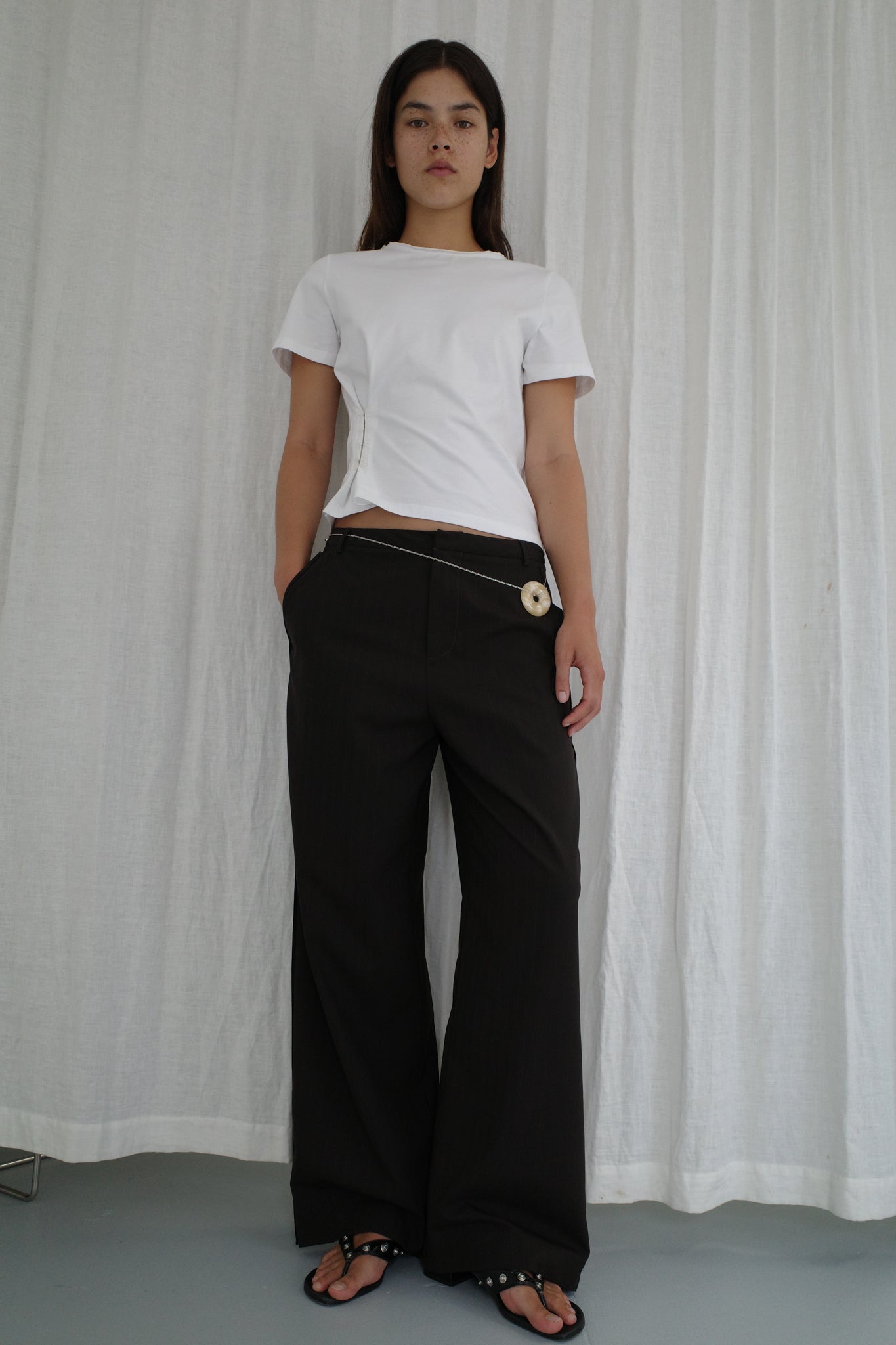 A model stands against a white draped backdrop wearing a fitted white short-sleeve t-shirt, Bind Pant - Charred Black Stripe by Deiji Studios with chain and disc waist details, and black embellished flip-flops.