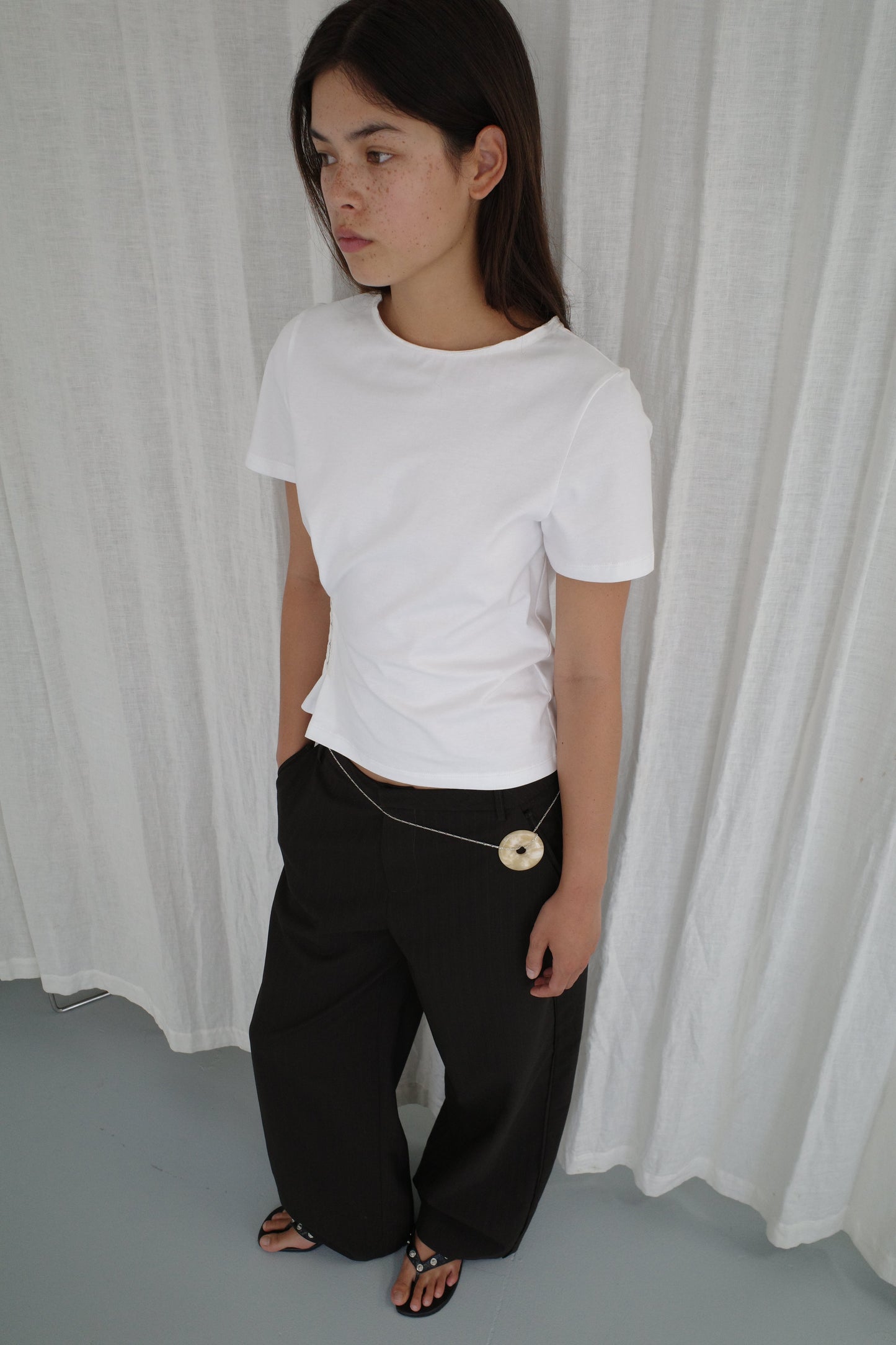 Model wearing the Pinch Tee - White from Deiji Studios, paired with wide-leg black trousers with a chain belt and round disc pendant, plus black strappy sandals, standing against a white linen curtain backdrop.