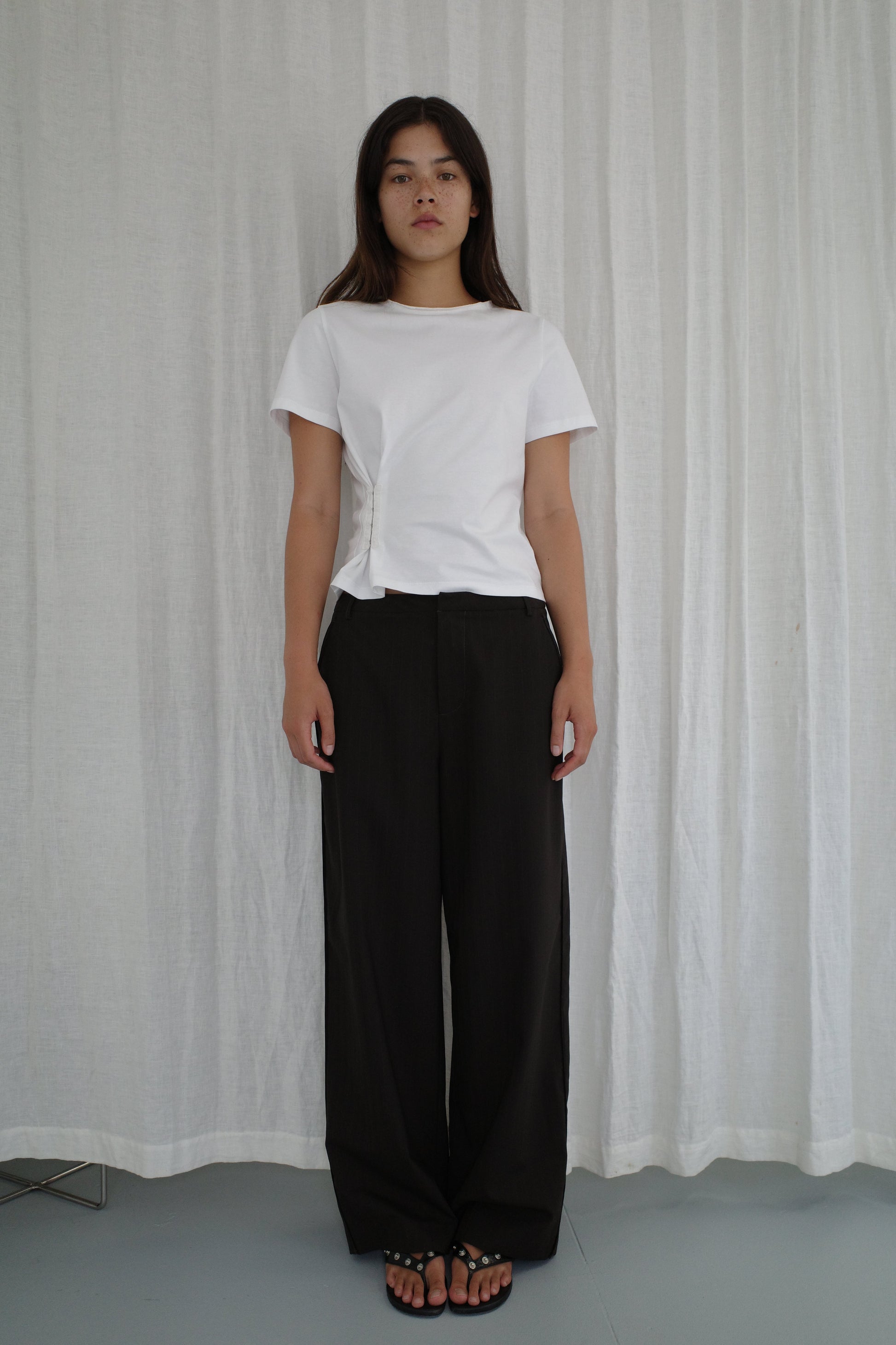 A woman with long dark hair stands in front of white curtains, wearing the Pinch Tee - White by Deiji Studios, loose black pants, and black sandals. She has a neutral expression and her arms are relaxed at her sides.