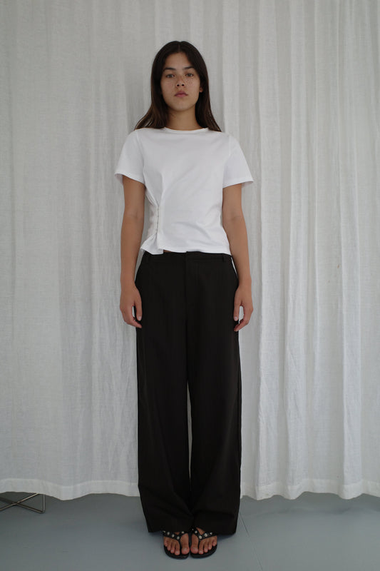 A woman with long brown hair and a neutral expression stands before white curtains, wearing the Pinch Tee - White from Deiji Studios, loose black pants, and black sandals. Her arms are relaxed at her sides.