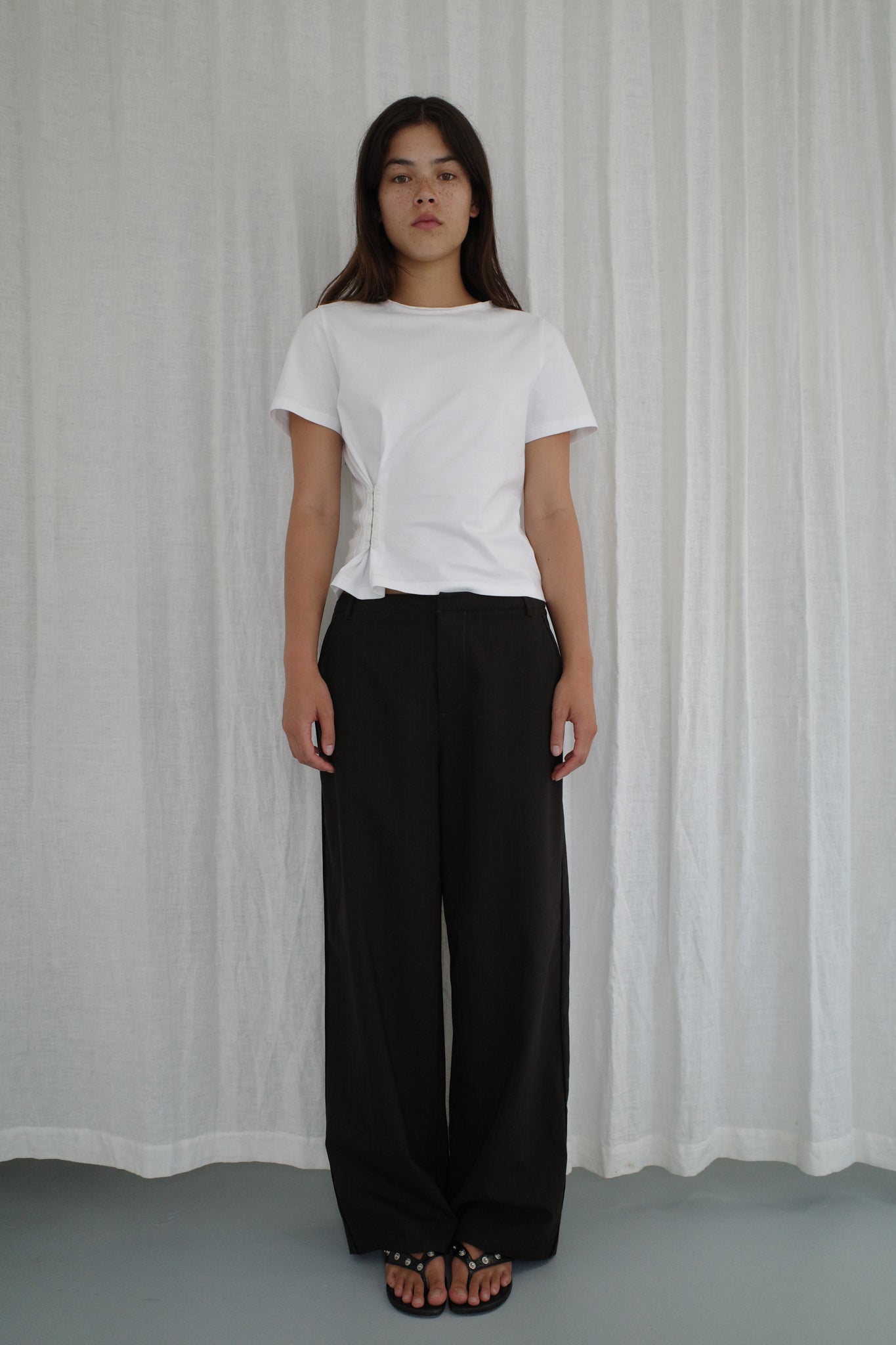 A woman stands before a white curtain, wearing a white short-sleeve t-shirt, the Bind Pant in Charred Black Stripe by Deiji Studios, and black sandals. She has a neutral expression with her arms relaxed at her sides.