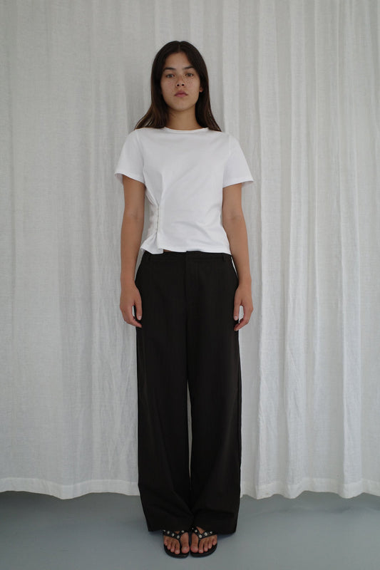 A woman with long brown hair stands before a white curtain, wearing a Deiji Studios white short-sleeve top, Bind Pant - Charred Black Stripe, and black sandals. She has a neutral expression.