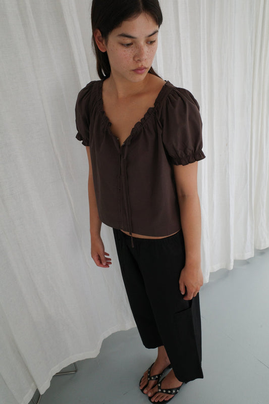 A person with shoulder-length dark hair stands against white curtains, wearing a Deiji Studios Close Shirt - Sepia, loose black pants, and black sandals with white spots, looking down and to the side.