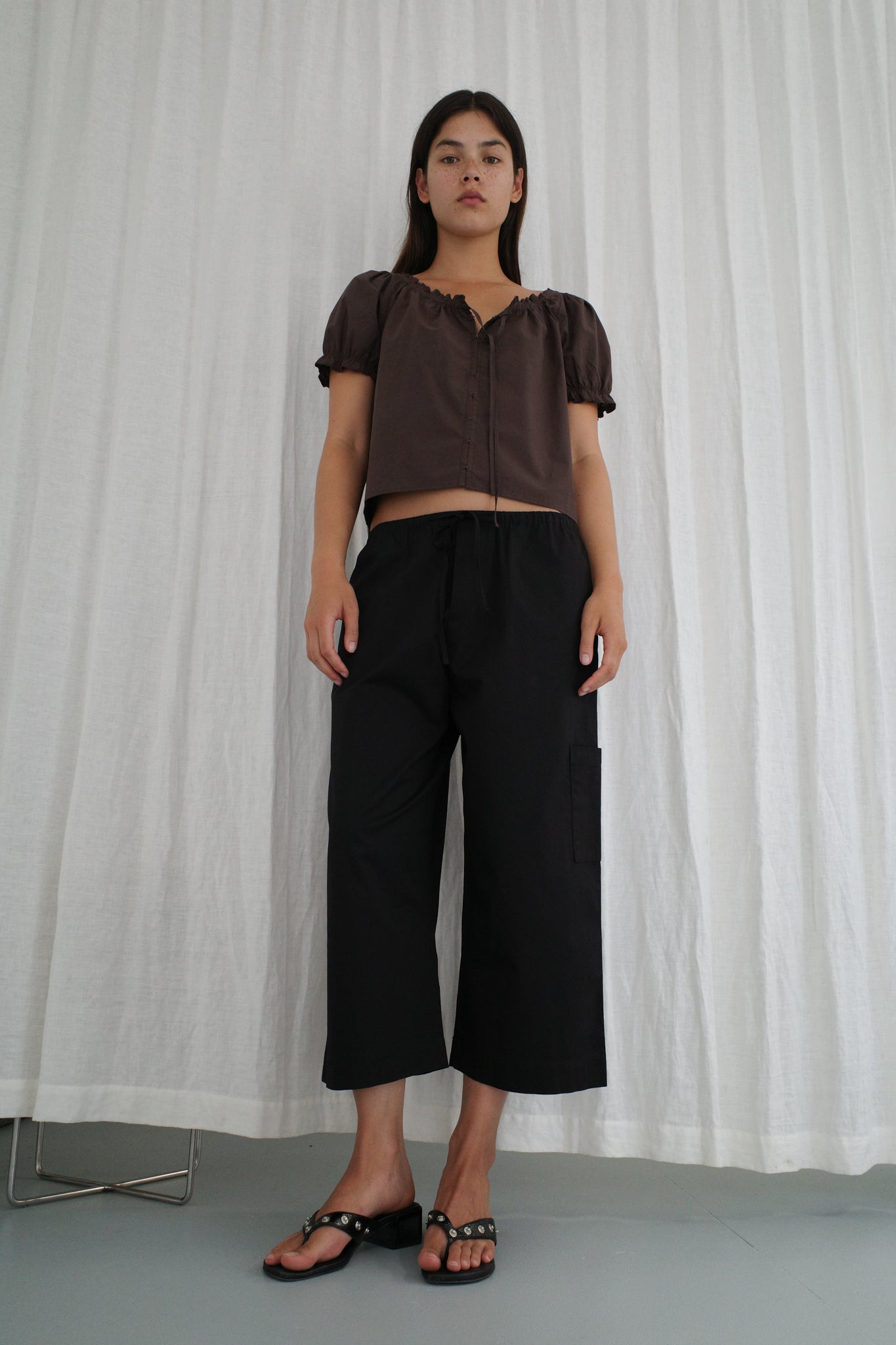 A person stands before a white curtain, wearing Deiji Studios' Close Shirt in Sepia, paired with loose black pants and black sandals. Their straight dark hair frames a neutral expression.