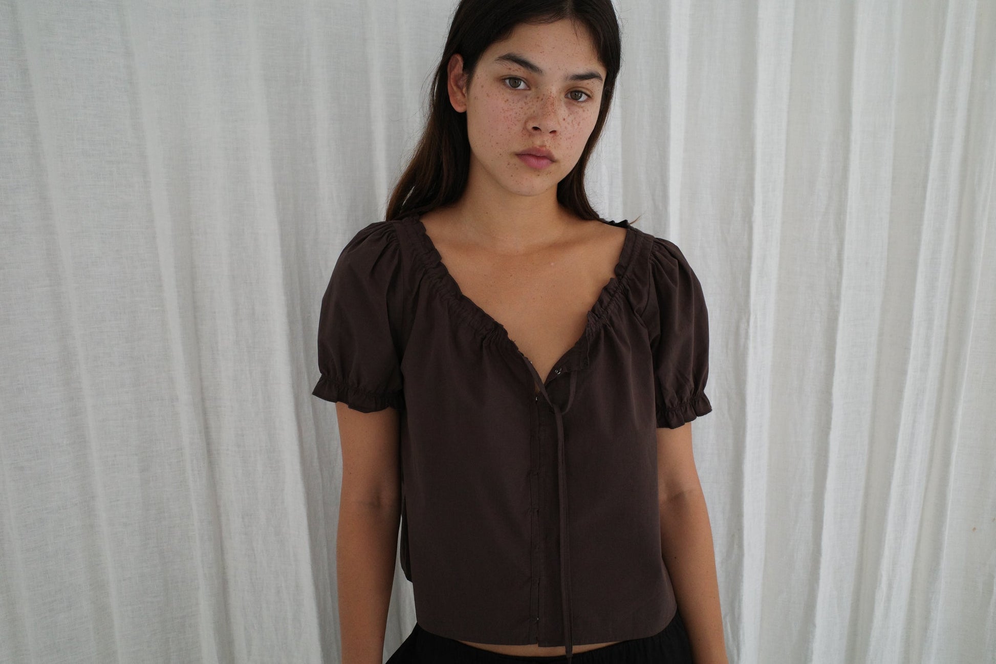 A woman with long dark hair and freckles stands before white curtains, wearing the Close Shirt - Sepia by Deiji Studios. She faces the camera with a neutral expression.