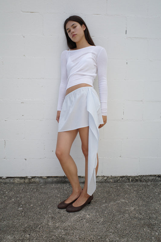 A woman with straight, dark hair and a neutral expression stands against a white brick wall, wearing the Deiji Studios Shoulder Knit Top in white, a light blue asymmetrical skirt with a draped panel, and brown flats.