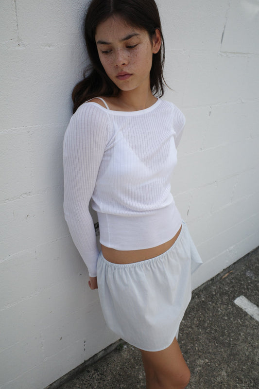 A woman with long dark hair stands against a white brick wall, wearing the Shoulder Knit Top - White from Deiji Studios and a light, flowy skirt. She looks down with a calm, thoughtful expression.